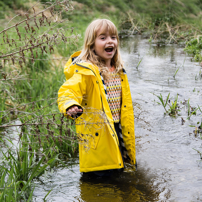 A blonde girl in a stream, wearing a Puddleflex Waterproof Fleece Lined Jacket in yellow. She's smiling and appears to be enjoying herself.